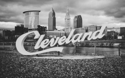 Black-and-white city skyline with a large cursive 'Cleveland' sign in the foreground by a rocky waterfront.