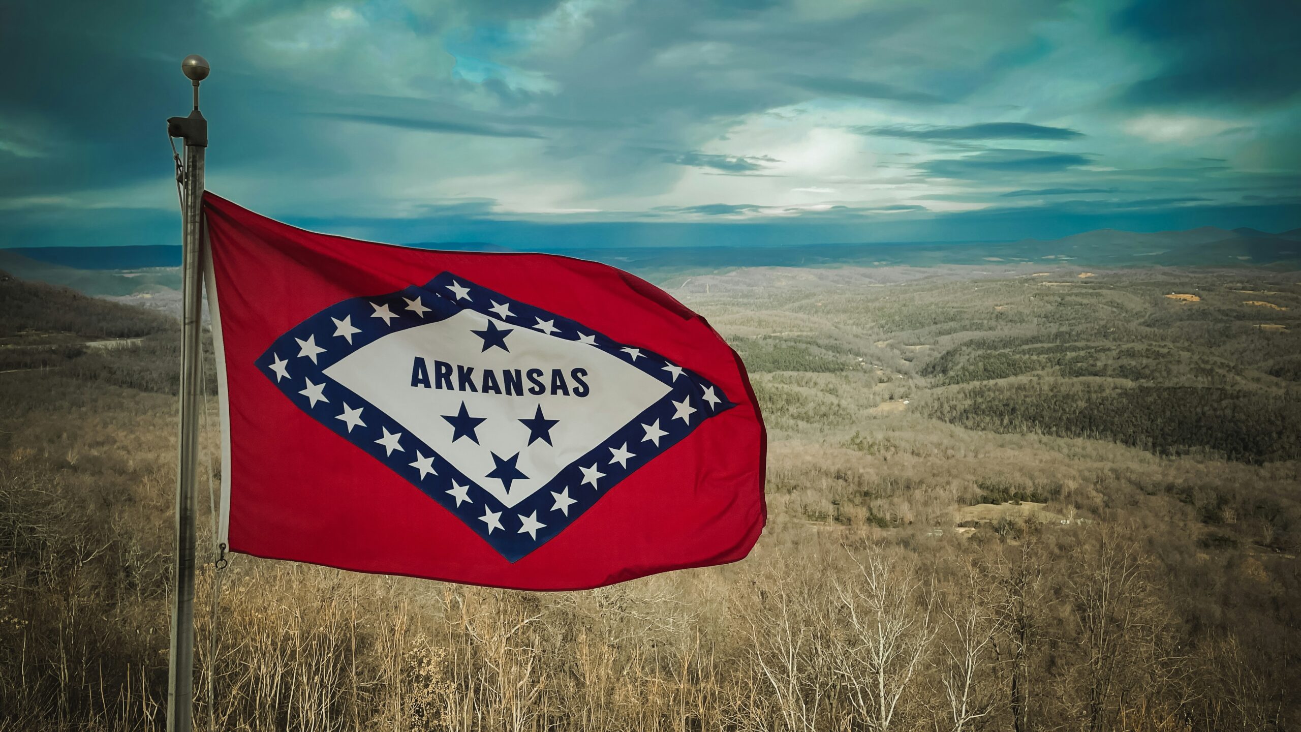 Texas AG Challenges ActBlue on Election Security Arkansas state flag fluttering on a pole with a distant hilly landscape in the background.”,